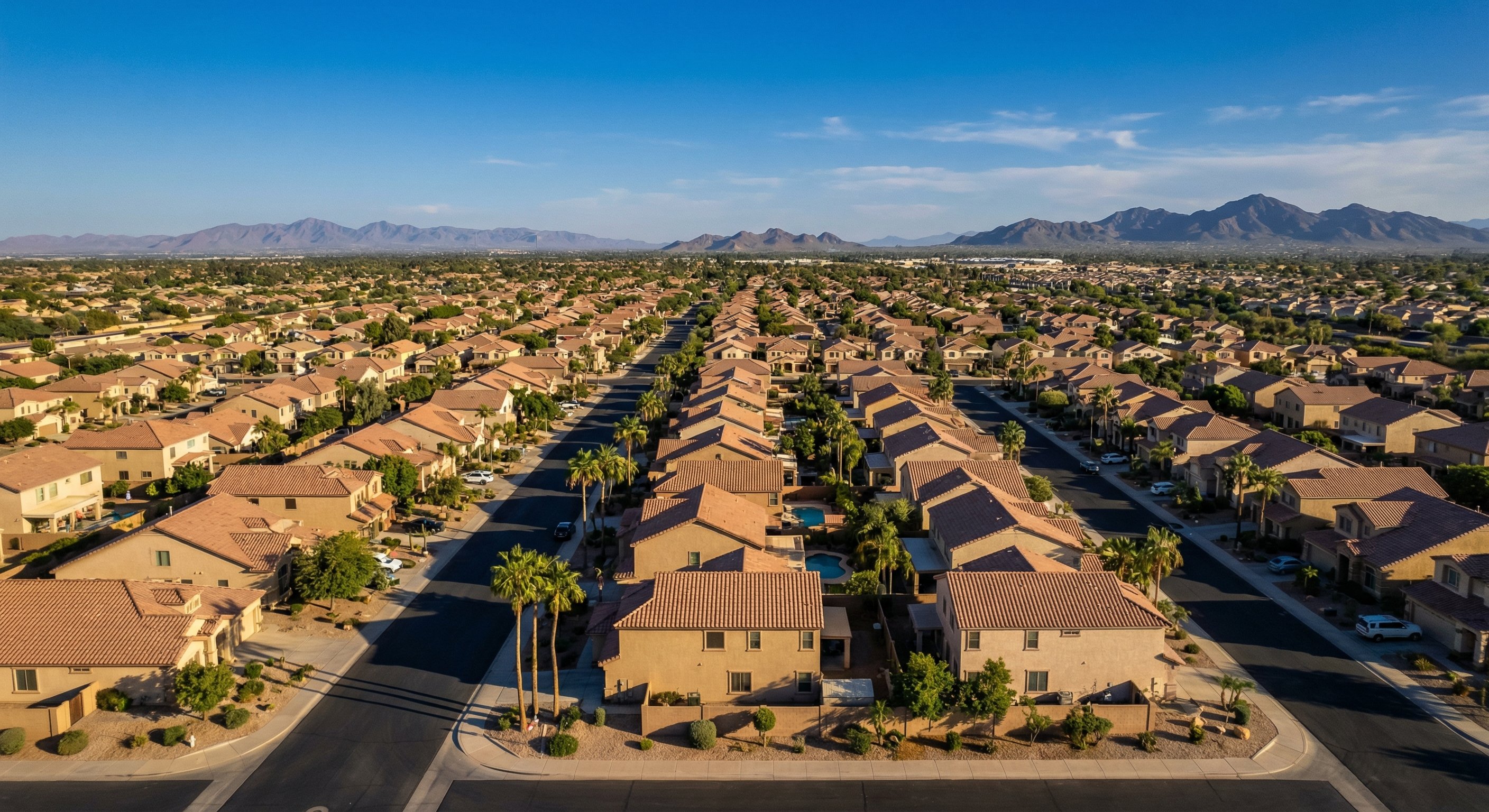 Aerial view of Phoenix metro subdivision with stucco homes, tile roofs, palm trees, and swimming pools visible beneath a deep blue desert sky