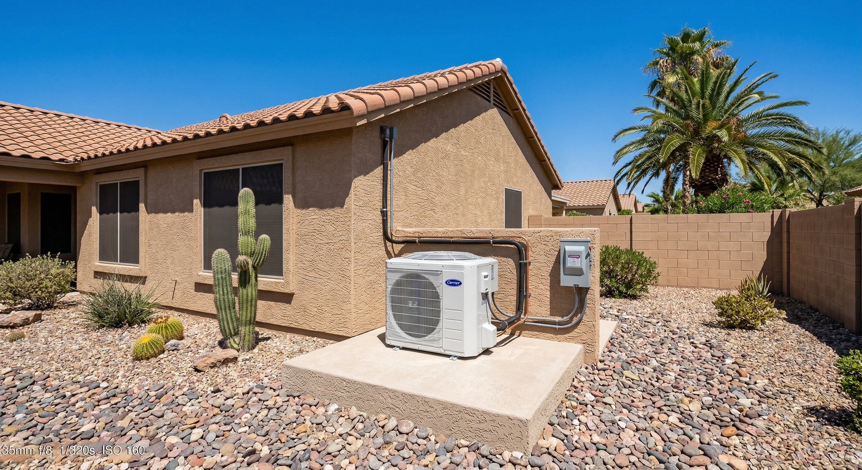 New Carrier AC condenser on concrete pad beside a tan stucco Arizona home with river rock landscaping, desert blue sky, saguaro cactus in background